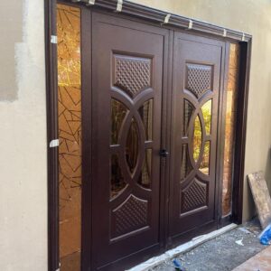 Ornate Double Glass DoorA stylish double door with dark wood finish, intricate geometric glass panels, and elegant carvings, currently under installation. Long Description: The Ornate Double Glass Door features a rich dark wood finish, complemented by intricate geometric patterns etched into the glass panels on both doors. The design includes detailed carvings with a lattice texture and oval accents, adding a touch of elegance and sophistication. The door appears to be in the process of installation, as evidenced by the protective tape along the frame and the surrounding construction debris. The adjacent tiled floor and overhanging greenery enhance theA stylish double door with dark wood finish, intricate geometric glass panels, and elegant carvings, currently under installation. Long Description: The Ornate Double Glass Door features a rich dark wood finish, complemented by intricate geometric patterns etched into the glass panels on both doors. The design includes detailed carvings with a lattice texture and oval accents, adding a touch of elegance and sophistication. The door appears to be in the process of installation, as evidenced by the protective tape along the frame and the surrounding construction debris. The adjacent tiled floor and overhanging greenery enhance the