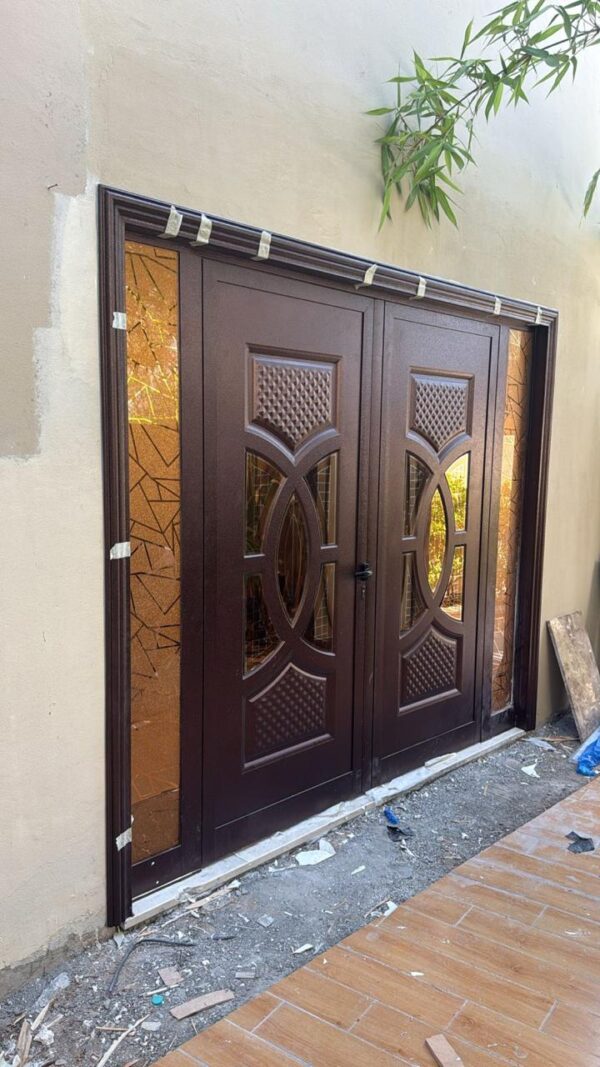 Ornate Double Glass DoorA stylish double door with dark wood finish, intricate geometric glass panels, and elegant carvings, currently under installation.  Long Description: The Ornate Double Glass Door features a rich dark wood finish, complemented by intricate geometric patterns etched into the glass panels on both doors. The design includes detailed carvings with a lattice texture and oval accents, adding a touch of elegance and sophistication. The door appears to be in the process of installation, as evidenced by the protective tape along the frame and the surrounding construction debris. The adjacent tiled floor and overhanging greenery enhance theA stylish double door with dark wood finish, intricate geometric glass panels, and elegant carvings, currently under installation.  Long Description: The Ornate Double Glass Door features a rich dark wood finish, complemented by intricate geometric patterns etched into the glass panels on both doors. The design includes detailed carvings with a lattice texture and oval accents, adding a touch of elegance and sophistication. The door appears to be in the process of installation, as evidenced by the protective tape along the frame and the surrounding construction debris. The adjacent tiled floor and overhanging greenery enhance the